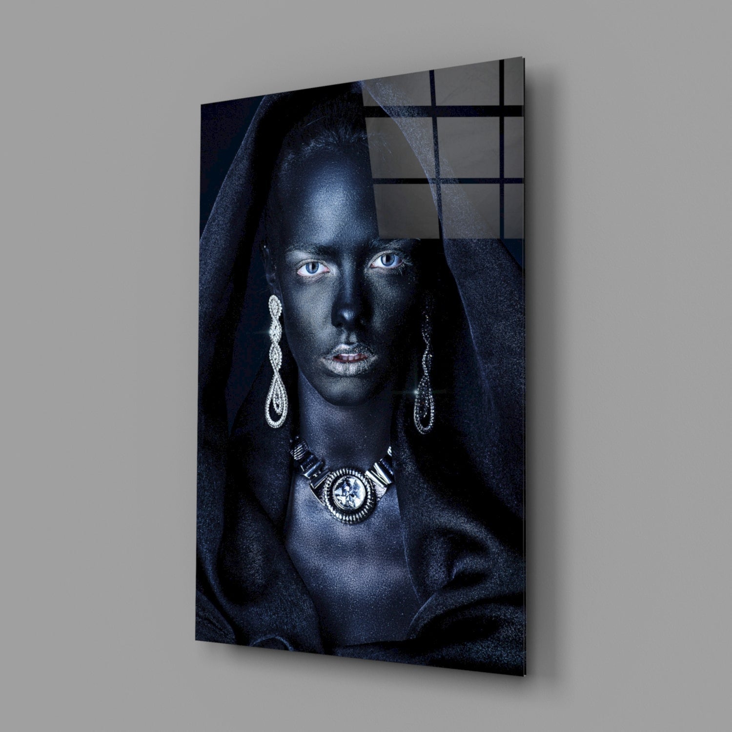 Glass wall art mounted on gray wall showing woman with dark skin, striking blue eyes, silver jewelry, and dark draped fabric against dark background.