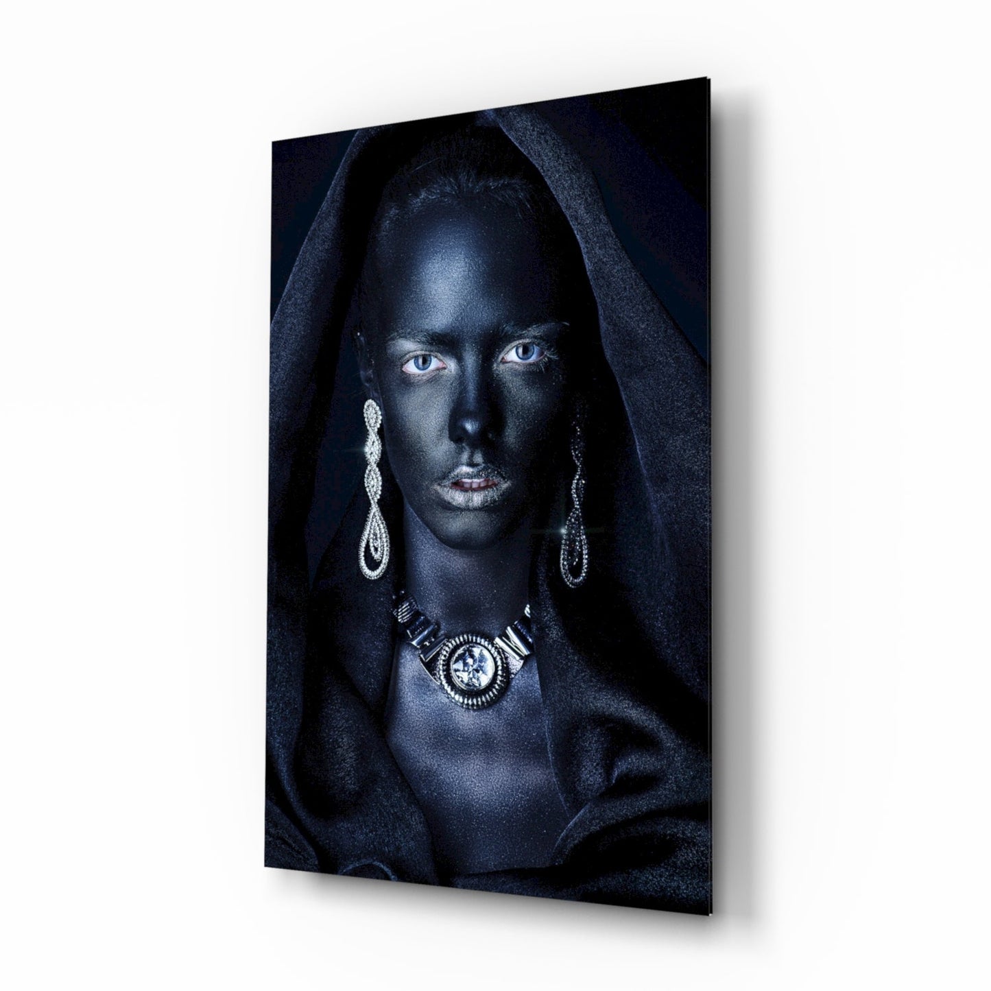 Woman with striking blue eyes wearing ornate silver jewelry and dark hooded fabric against black background on glass wall art, angled perspective view.