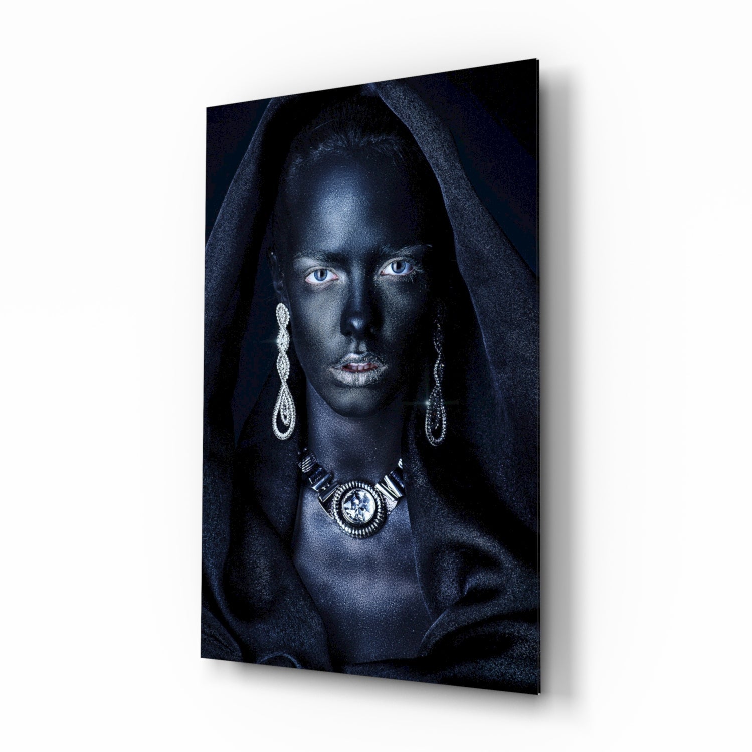 Woman with striking blue eyes wearing ornate silver jewelry and dark hooded fabric against black background on glass wall art, angled perspective view.