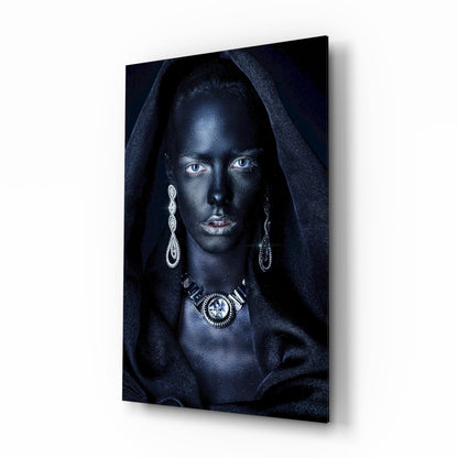 Woman with striking blue eyes wearing ornate silver jewelry and dark hooded fabric against black background on glass wall art, angled perspective view.