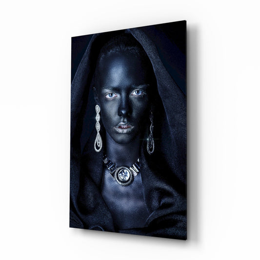 Woman with striking blue eyes wearing ornate silver jewelry and dark hooded fabric against black background on glass wall art, angled perspective view.