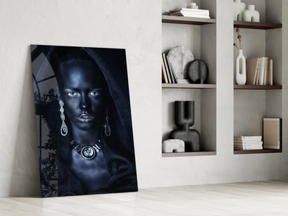 Glass wall art featuring a blue-toned portrait of a woman with striking eyes and ornate jewelry, leaning against white wall beside shelving.