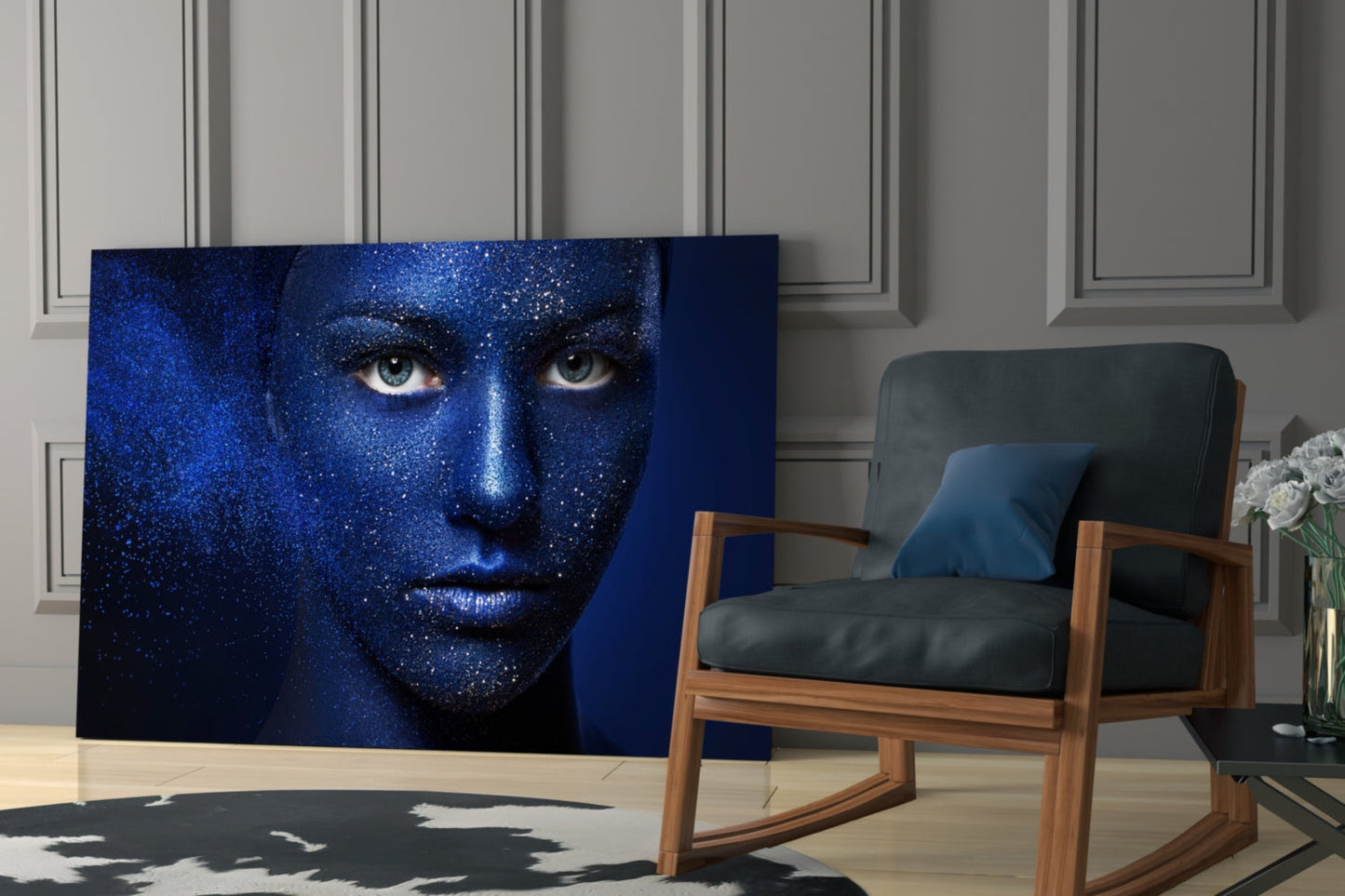 Blue glitter-textured woman's face portrait displayed as glass wall art in modern living room with wooden chair and white flowers.