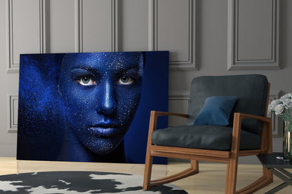Blue glitter-textured woman's face portrait displayed as glass wall art in modern living room with wooden chair and white flowers.
