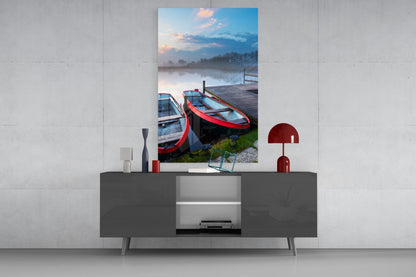 Glass wall art depicting red and blue boats moored at a wooden dock beside a misty lake at sunrise, displayed above a gray media console.