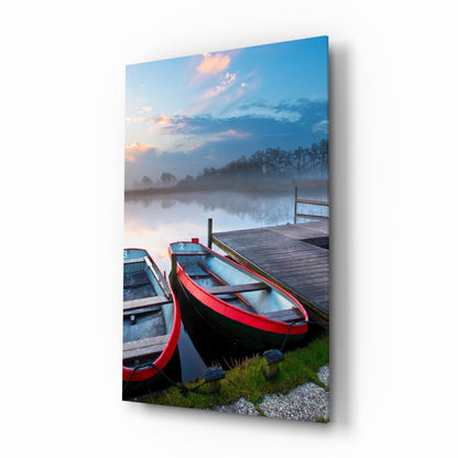 Red and black boats moored at wooden dock with misty lake, trees, and sunset sky reflected in still water.