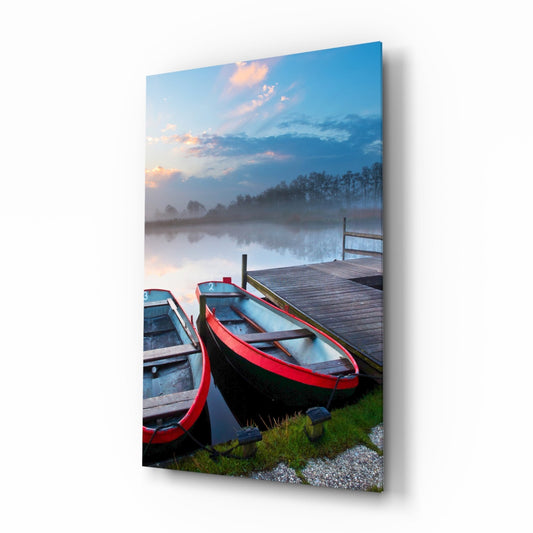 Red and black boats moored at wooden dock with misty lake, trees, and sunset sky reflected in still water.