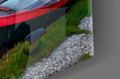 Close-up of red boat hull with gray weathered wood beam, green grass, and white pebble beach along waterside.