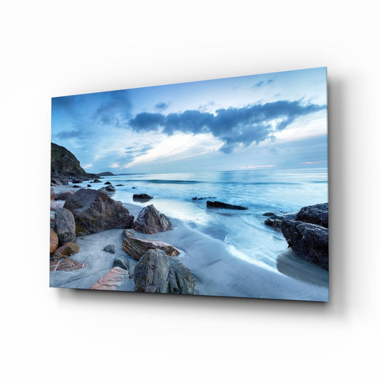 Angled view of glass wall art displaying a rocky coastline at dusk with blue-toned ocean, beach, and cloudy sky.