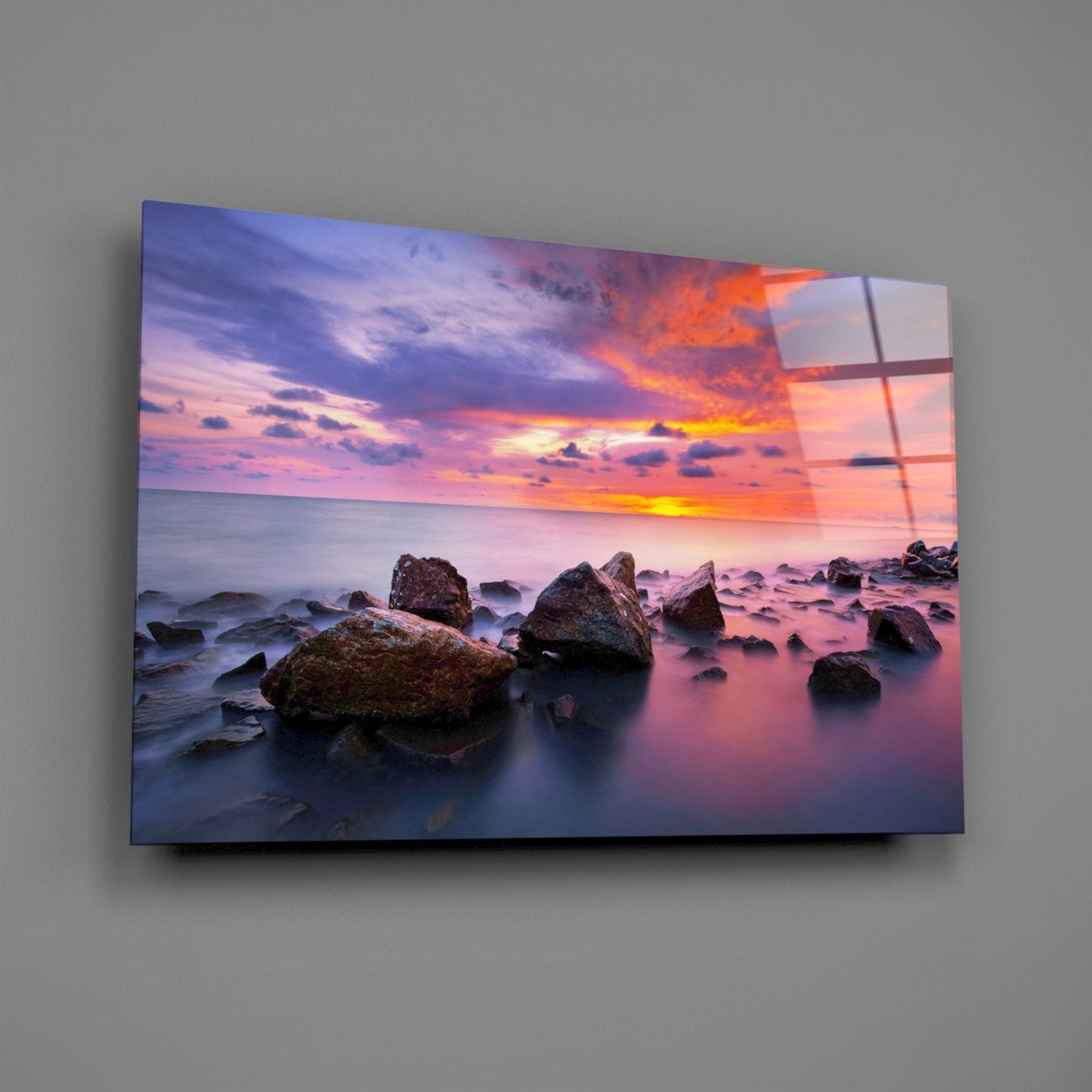 Glass wall art mounted on gray wall displaying rocky seashore at sunset with pink and orange sky.