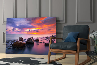 Glass wall art displaying a vibrant sunset seascape with rocky shore displayed in modern living room setting.