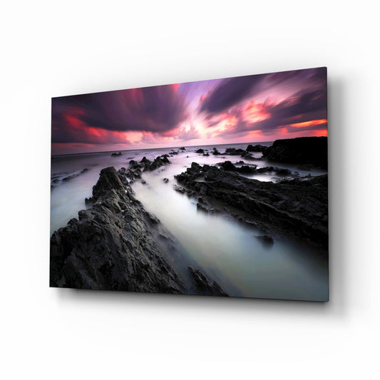 Glass wall art displayed at an angle showing a rocky coastal sunset with crimson sky and misty white water.