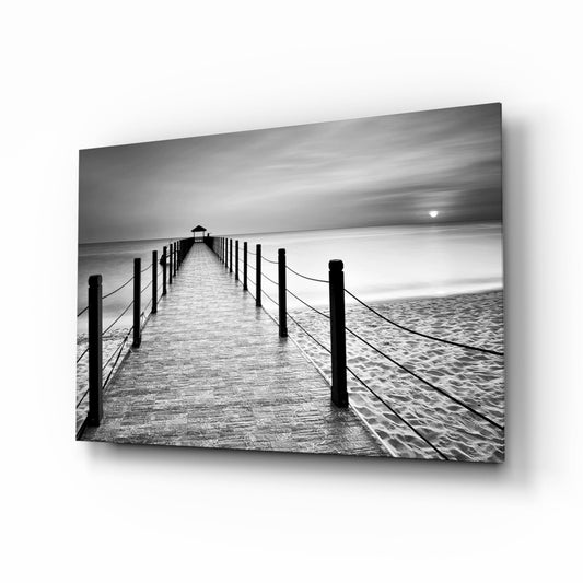 Black and white photograph of a wooden pier with rope railings extending toward the ocean and a small structure at the end, mounted on glass with a drop shadow effect.