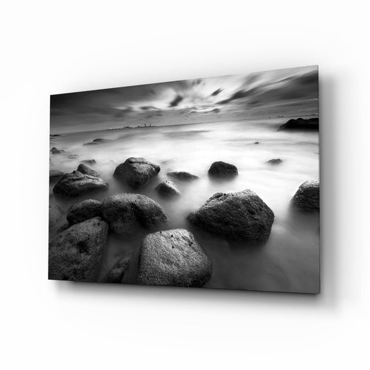 Black and white glass wall art showing rocky beach with misty waves and dramatic cloudy sky, displayed at an angle.