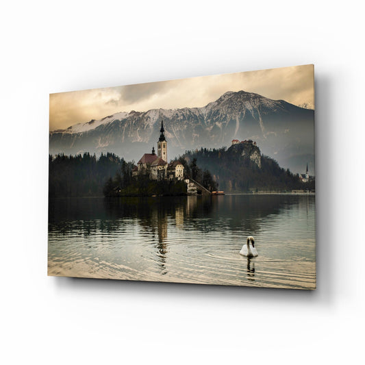 Glass wall art displaying a serene alpine lake scene with a church on an island, snow-capped mountains, a swan, and forested shoreline in warm golden tones.