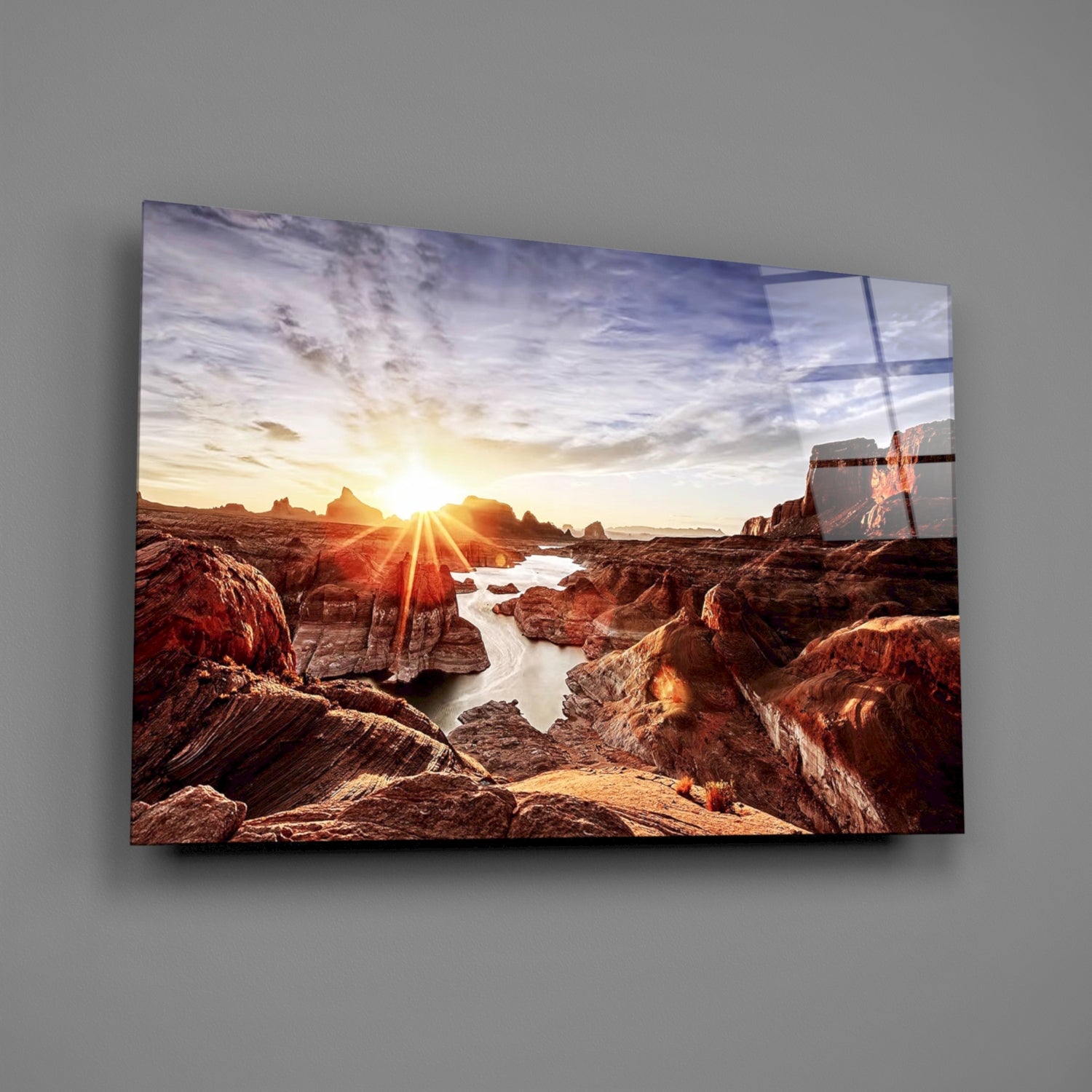 Glass wall art displaying a rocky canyon landscape at sunset with golden light, water stream, and a sailboat on the right side, mounted on a gray wall.