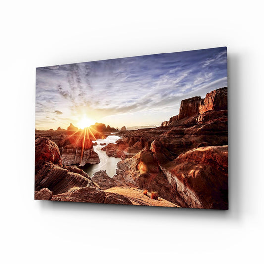 Glass wall art displaying a rocky canyon landscape at sunset with golden light, water channels, and red stone formations under a cloudy sky.