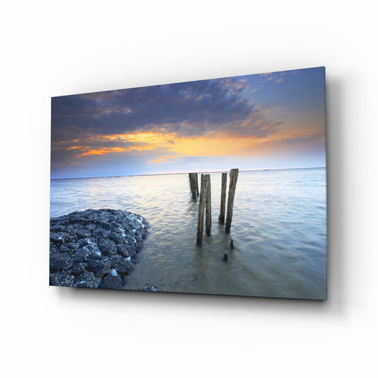 Weathered wooden pier posts extending into calm blue water at golden sunset, with rocky shore visible on left side.