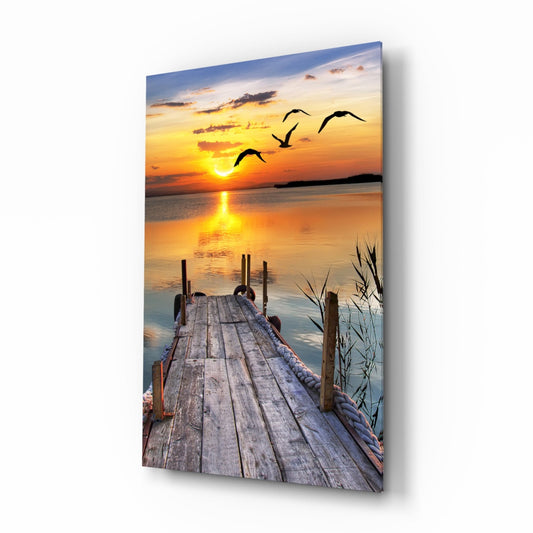 Wooden dock extending into calm water at sunset with silhouetted birds flying overhead and golden sun reflecting on the water's surface.