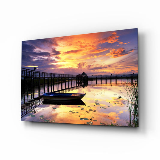 A wooden rowboat floats in shallow water beside a wooden pier at sunset, with golden and orange clouds reflected in the still water.