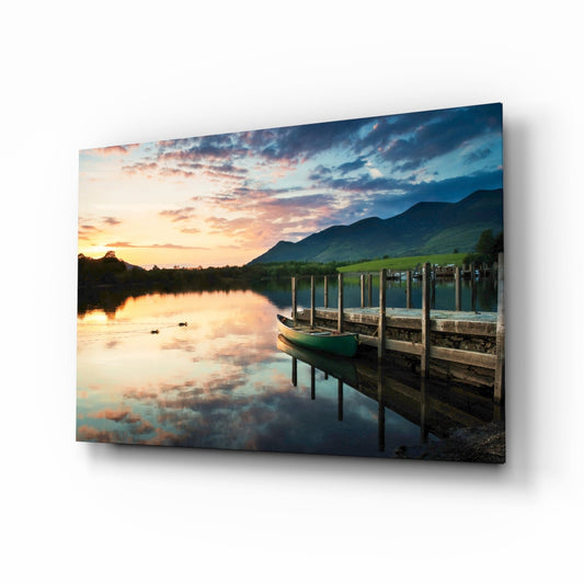 Green canoe moored at wooden dock at sunset with mountains, water reflection, and colorful sky.