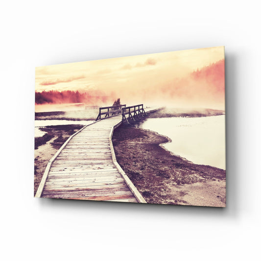 A wooden boardwalk curves through misty wetlands at sunrise, with evergreen trees and calm water visible under warm peachy-pink skies.