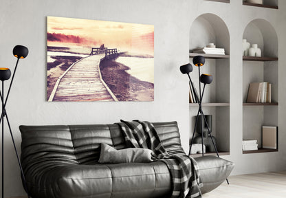 Glass wall art print of a wooden boardwalk extending toward misty water and trees in sepia tones, displayed above a black leather sofa.