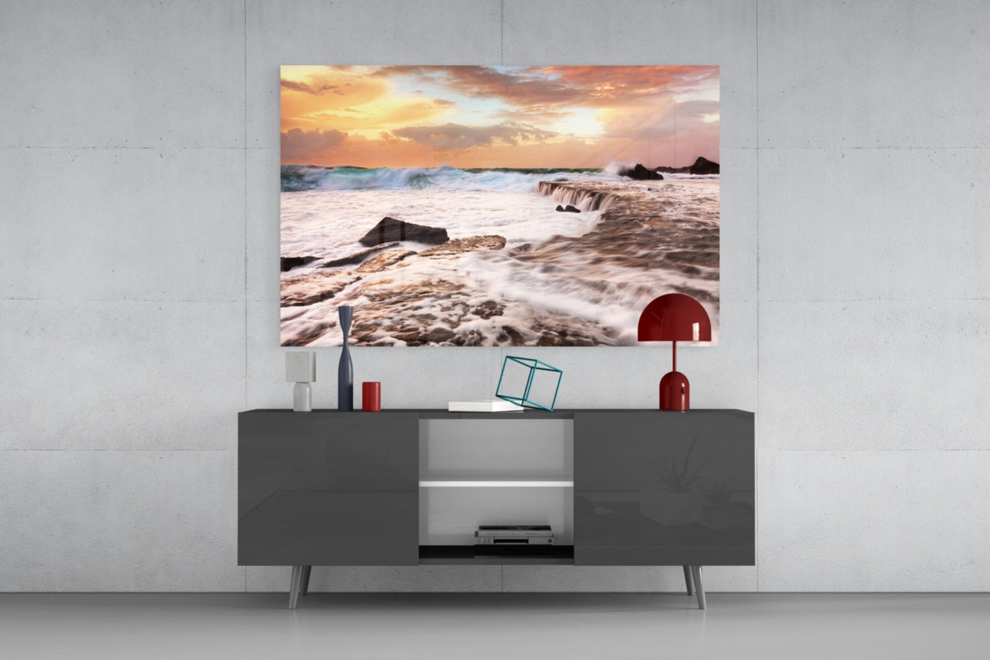 Vicious Waves Glass Wall Art depicting a dramatic sunset beach scene with golden sky, crashing waves, and dark rocks, displayed above a gray media console.