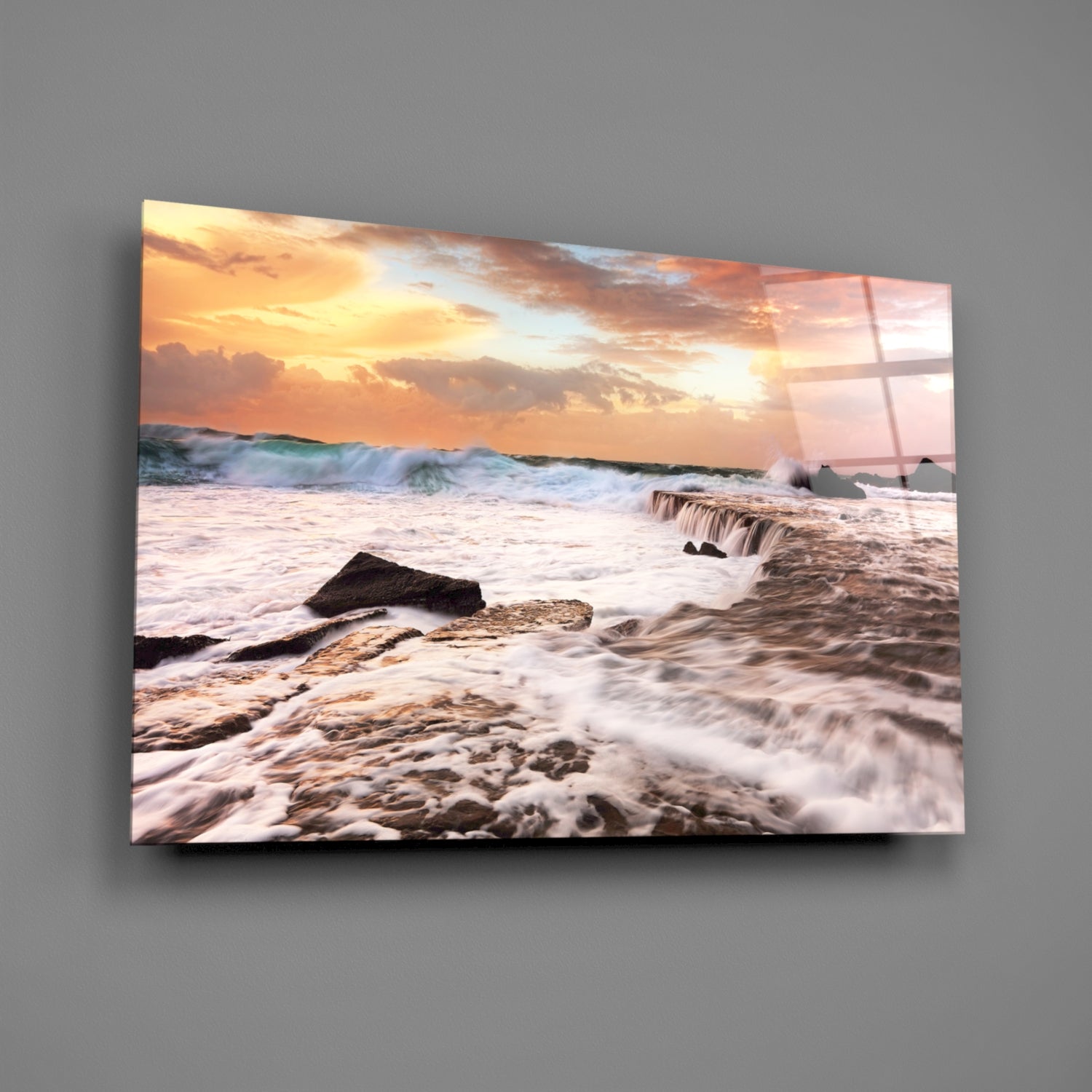 Coastal sunset scene with crashing waves and rocky shoreline printed on glass panel mounted on gray wall.