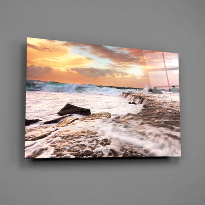 Coastal sunset scene with crashing waves and rocky shoreline printed on glass panel mounted on gray wall.
