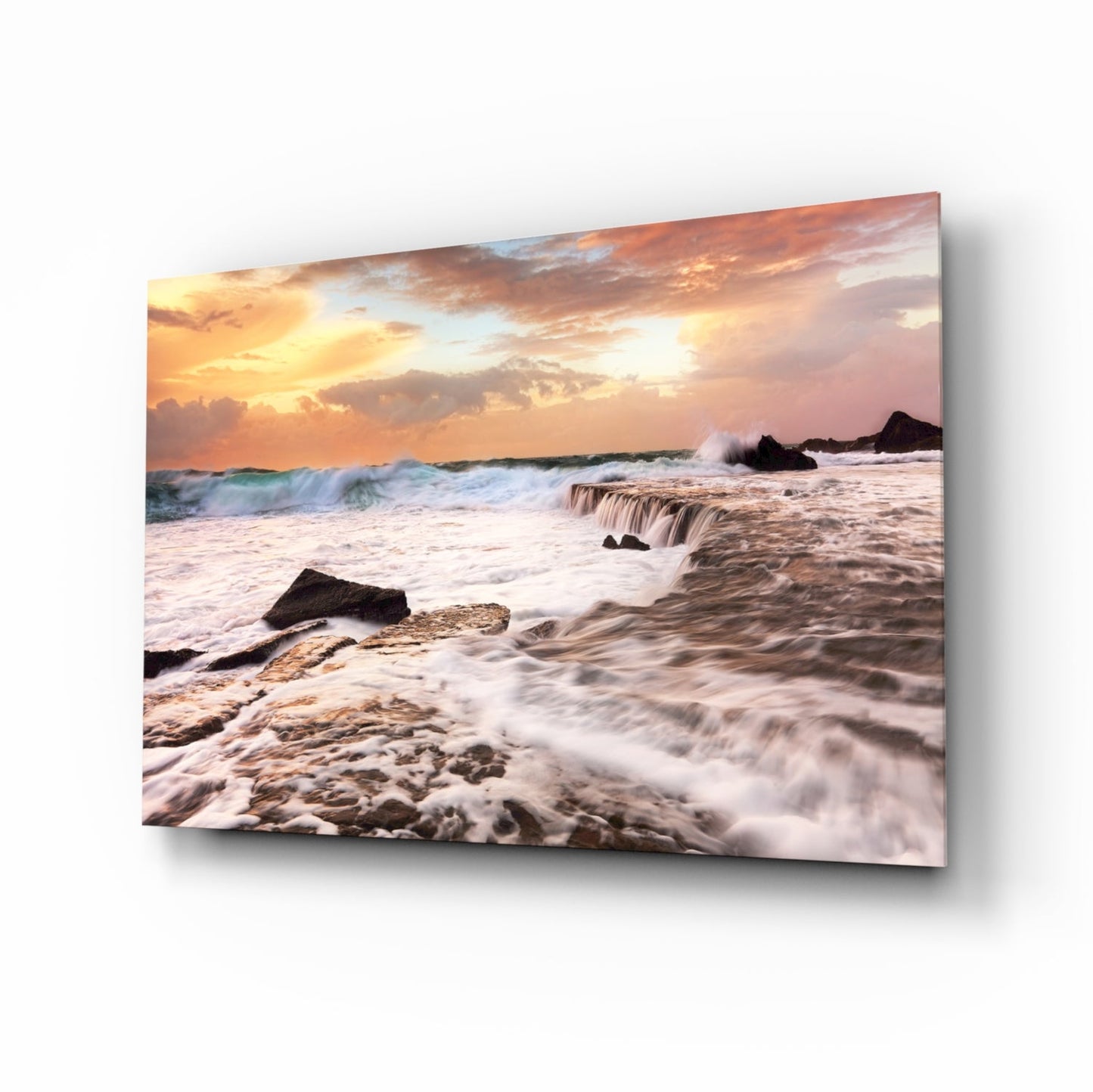 Coastal sunset scene with rolling waves, rocky shore, and golden-orange sky displayed on tilted glass wall art panel.