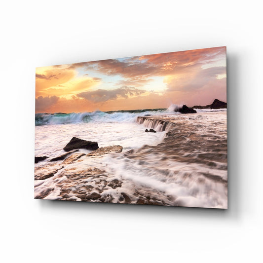 Coastal sunset scene with rolling waves, rocky shore, and golden-orange sky displayed on tilted glass wall art panel.