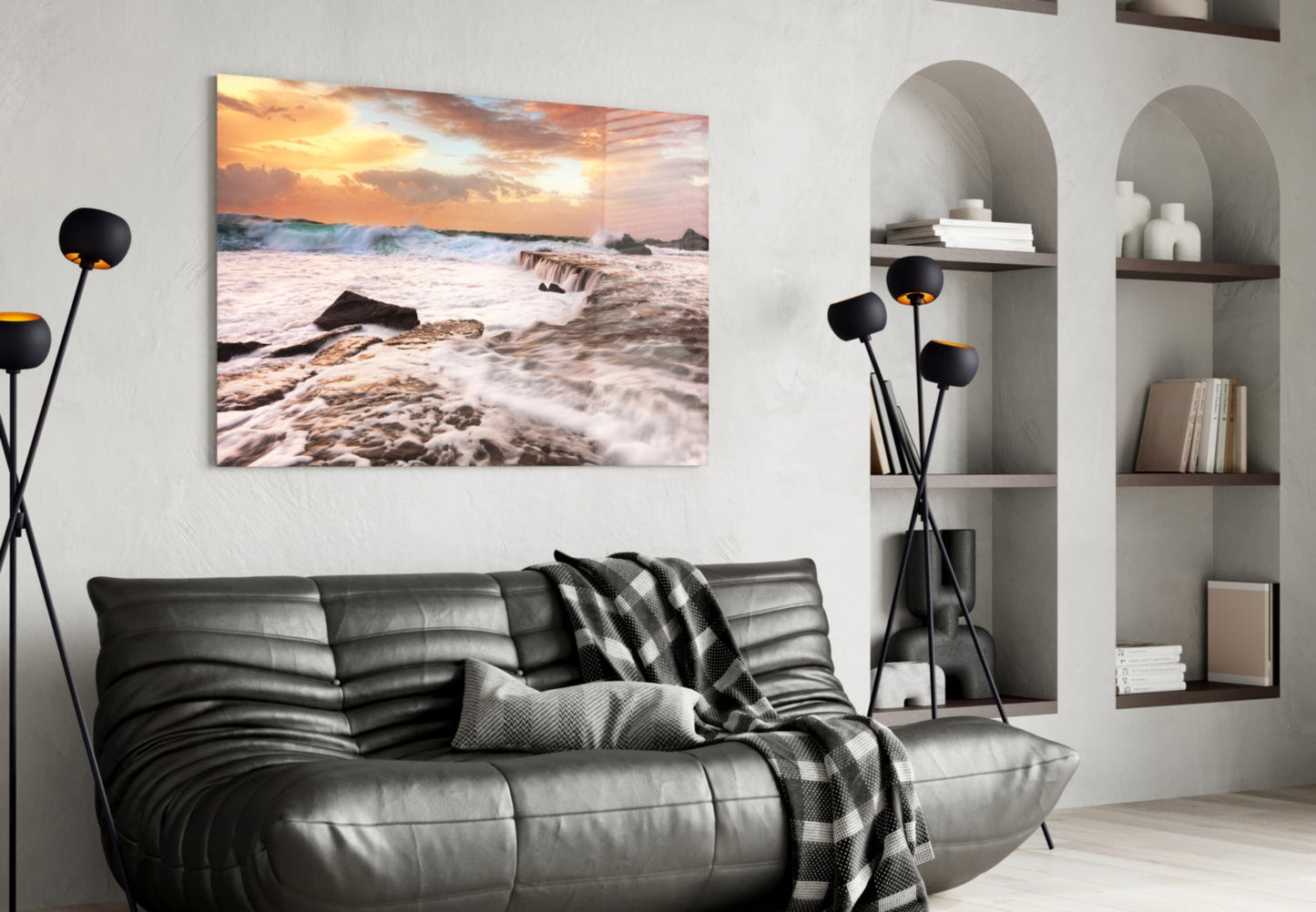 Vicious Waves Glass Wall Art depicting a sunset beach scene with golden sky and crashing waves displayed above a black leather sofa in a modern living room.
