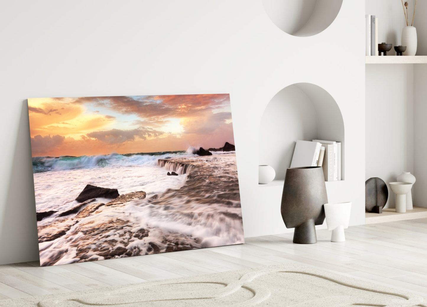 Vicious Waves Glass Wall Art leaning against white wall showing coastal sunset with golden sky, crashing waves, and dark rocks.