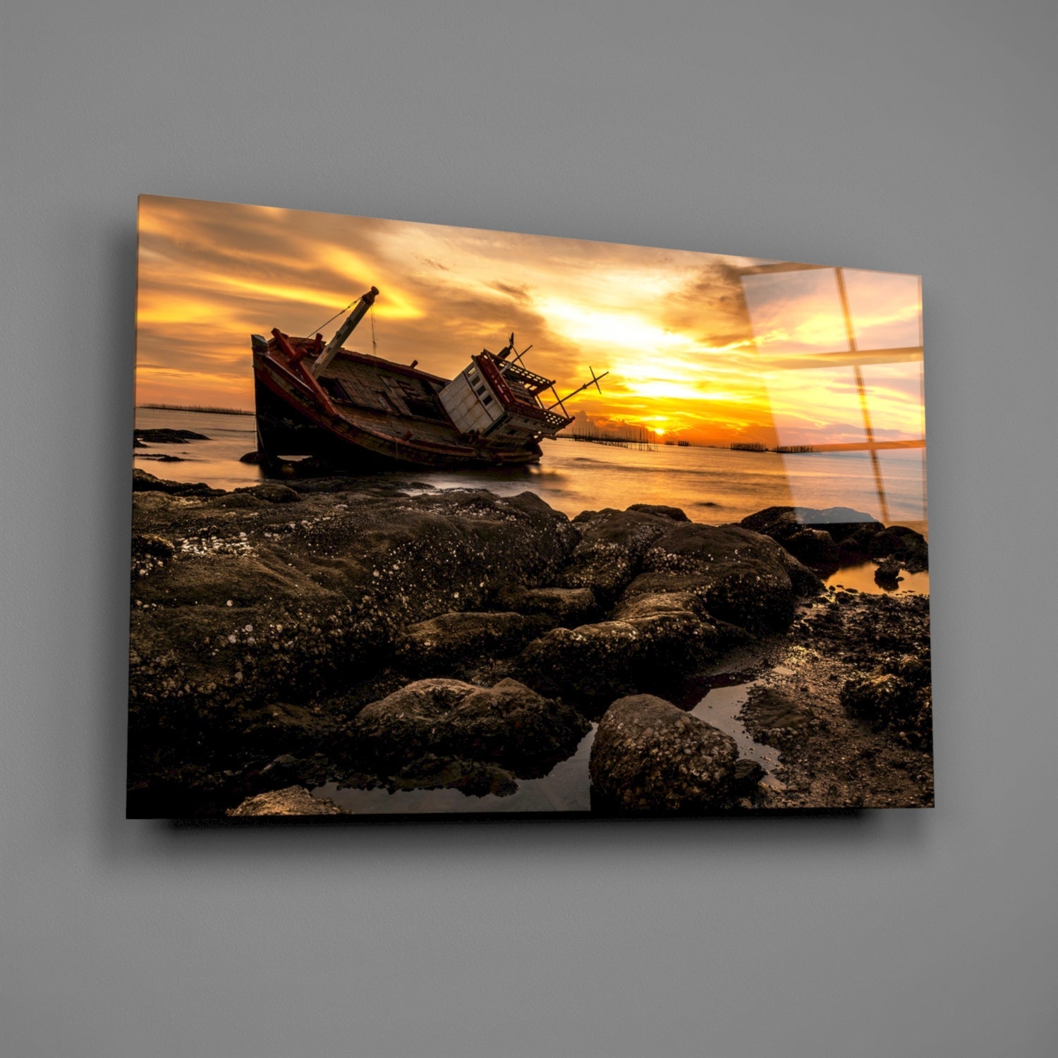 Angled front view of glass wall art depicting a wrecked boat on rocky shore at golden sunset, displayed against gray background.