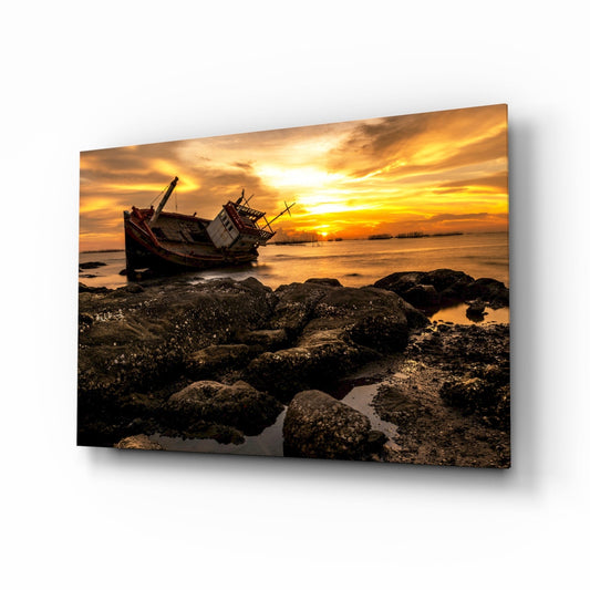 Shipwreck glass wall art displaying a rusted tilted boat at sunset with golden sky, rocky foreground, and calm water.