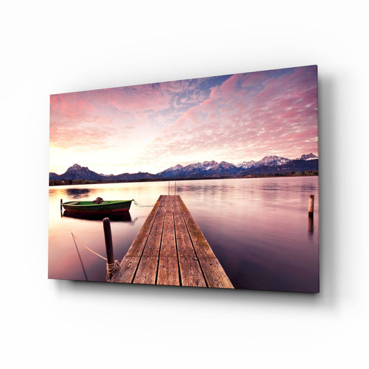 Wooden dock extending into calm lake with green boat, snow-capped mountains, and pink sunset sky on glass wall art panel.