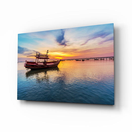 A glass wall art panel displaying a red wooden fishing boat on calm water at sunset, with pink and gold sky reflections, mounted at an angle showing dimensional depth.
