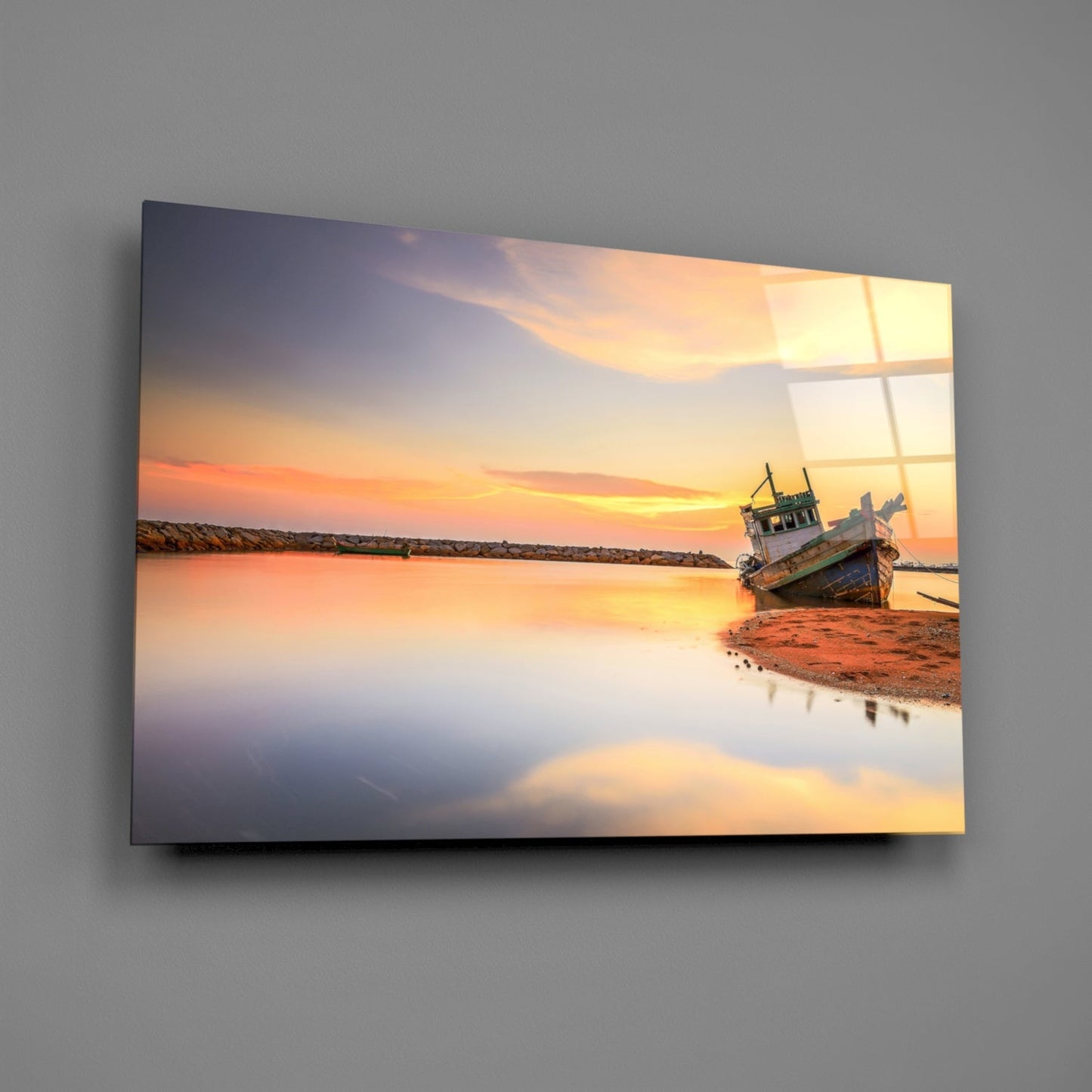 Glass wall art mounted on gray wall displaying a sunset coastal scene with a beached fishing boat reflected in still water.