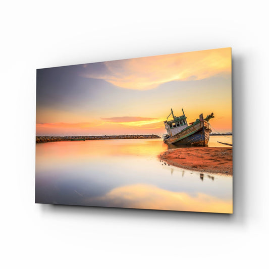 Glass wall art print displaying an abandoned wooden fishing boat beached at sunset with golden sky reflection in shallow water.