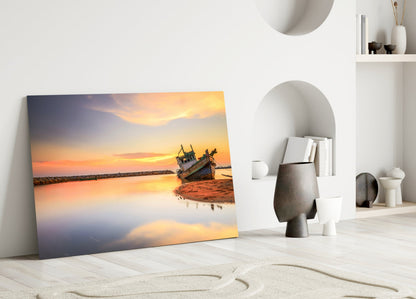 Glass wall art leaning against white wall displays golden sunset over calm water with beached wooden boat.