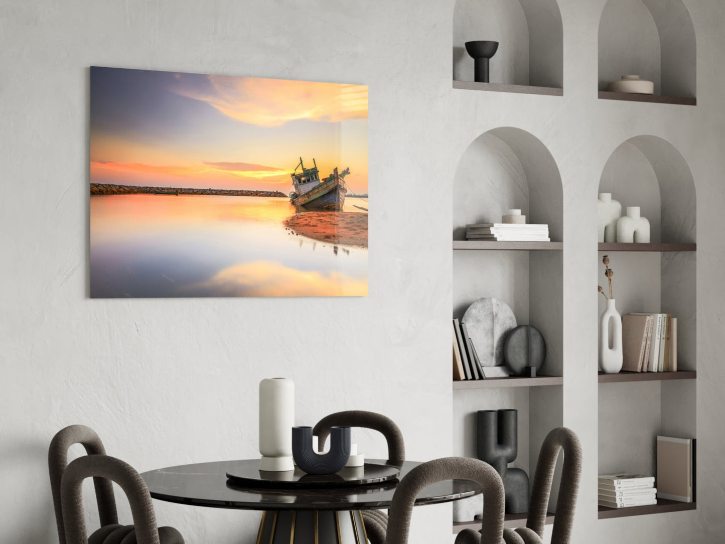 Glass wall art depicting a golden sunset over calm water with a moored fishing boat, displayed above a black dining table in a modern interior.