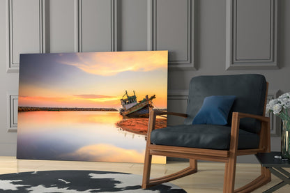 Glass wall art displaying a wooden fishing boat at sunset on a beach, displayed in a modern living room next to a wooden chair.