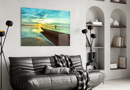 Glass wall art depicting a wooden kayak beached at sunset by calm turquoise water, displayed above a black leather sofa in a modern living room.