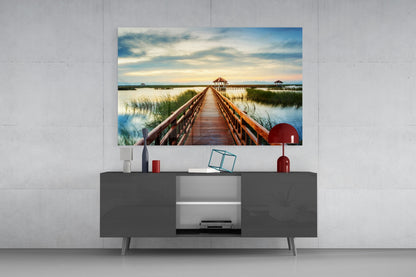 Glass wall art displaying a wooden dock extending into a wetland landscape with pavilions, mounted above a dark gray media console with red decorative accessories.