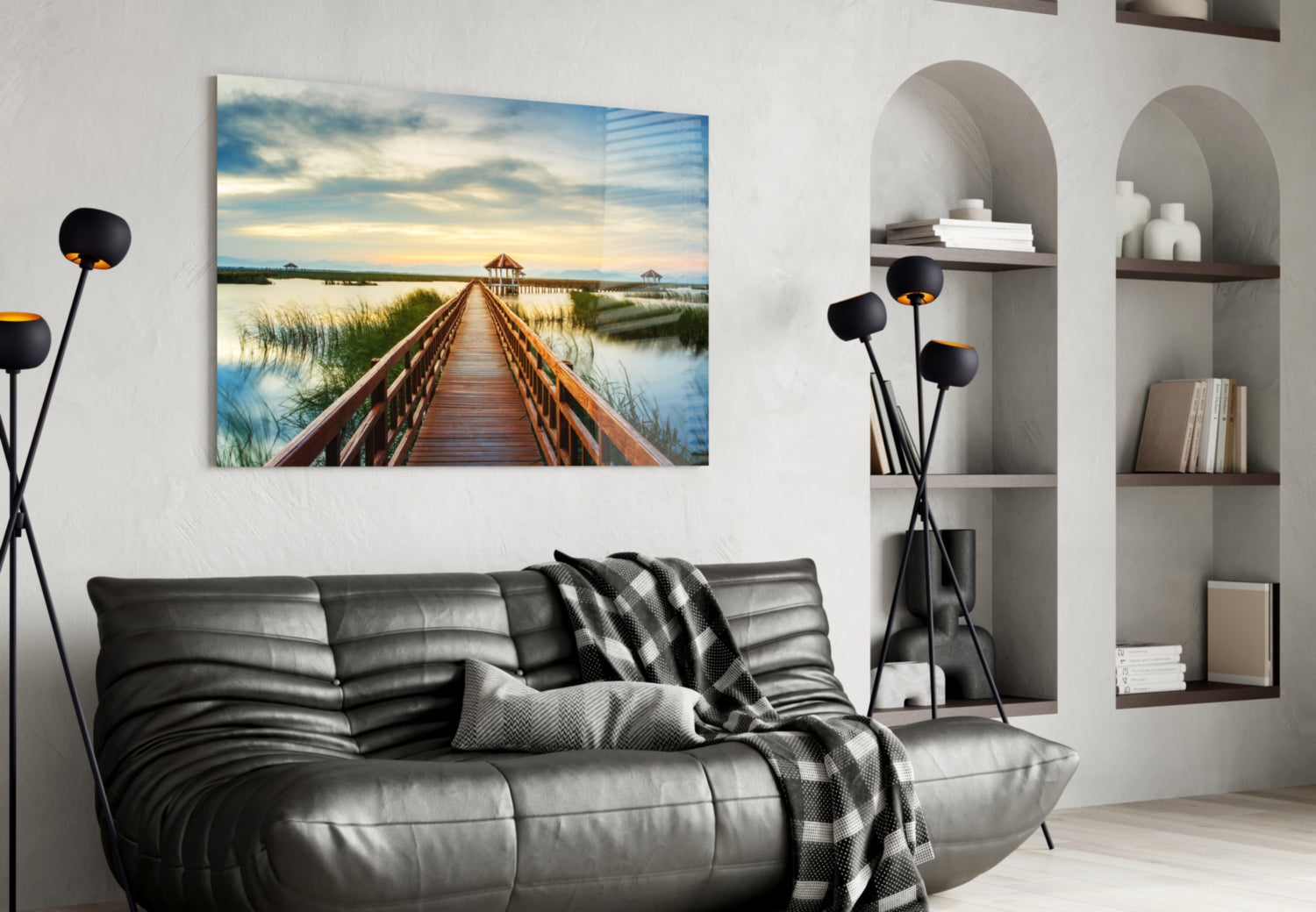 Glass wall art depicting a wooden boardwalk extending toward a gazebo across calm water at sunset, displayed above a gray sofa in a modern living room.