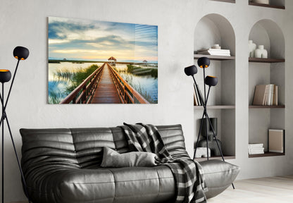 Glass wall art depicting a wooden boardwalk extending toward a gazebo across calm water at sunset, displayed above a gray sofa in a modern living room.