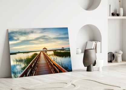 Glass wall art leaning against white wall displays wooden dock extending into calm lake under blue sky with clouds.