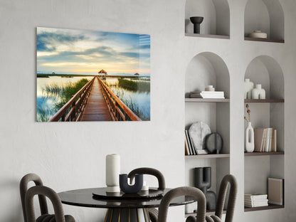 Glass wall art depicting a wooden boardwalk extending toward gazebos across calm blue water under a cloudy sky.