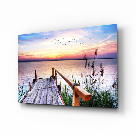 Wooden dock with railing extending toward pastel sunset sky, reeds along water's edge, birds flying overhead, displayed as glass wall art.